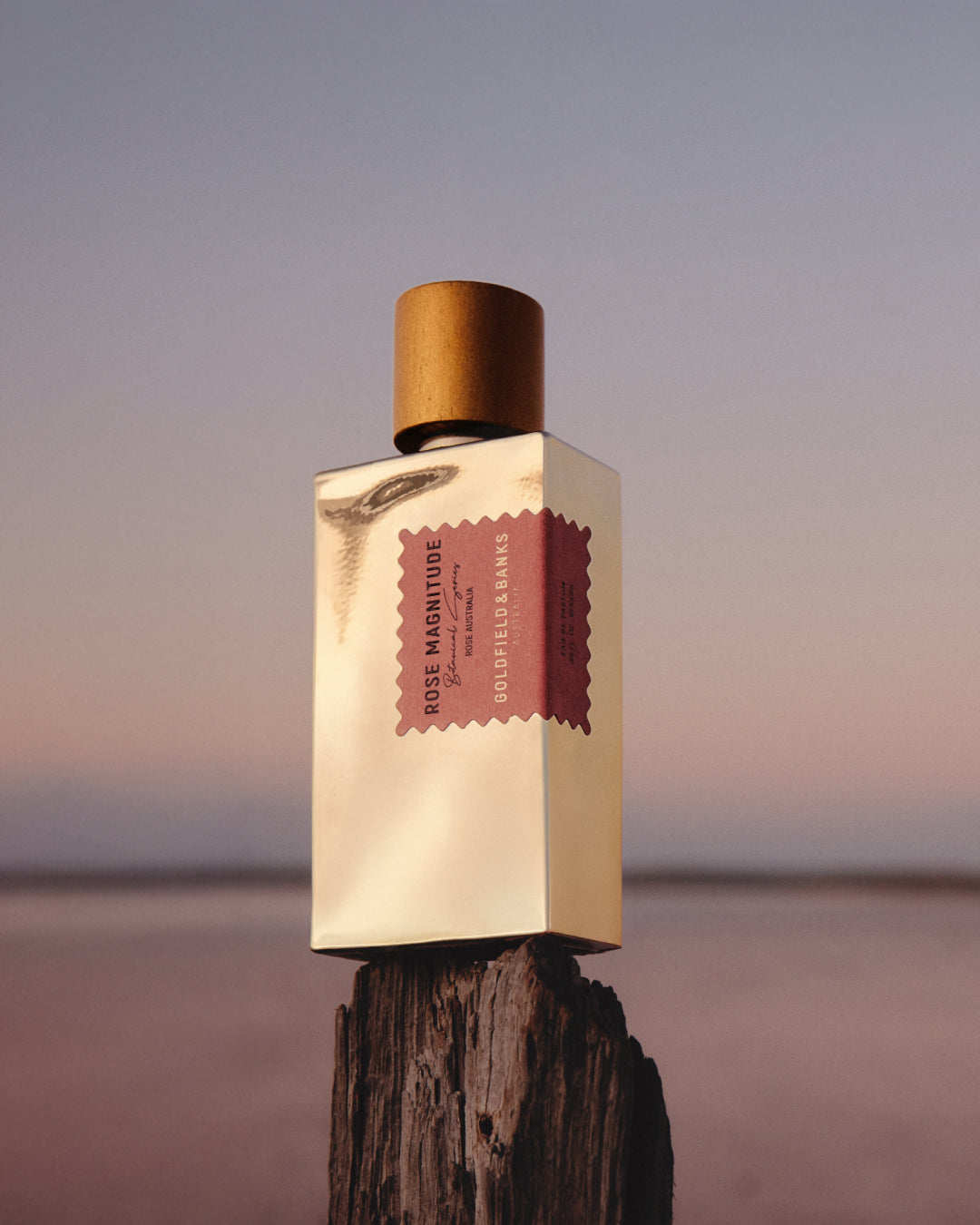 Luxury perfume bottle displayed on a wooden post against a soft sunset sky, featuring a minimalist gold rectangular bottle with a wooden cap and a pink label reading “Rose Magnitude.”