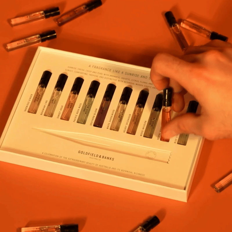 Flat lay of a luxury perfume discovery set with multiple fragrance samples arranged in a presentation box on an orange background