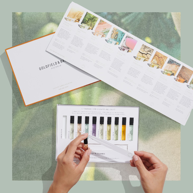 Hands holding a luxury fragrance sample kit with multiple perfume vials and a product catalog laid out on a textured surface.